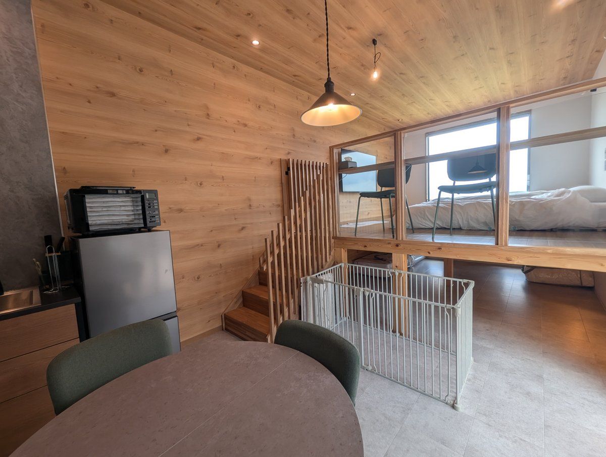Un interior moderno con techo de madera, un comedor con mesa y sillas, una cocina con refrigerador y una cama en una habitación con ventana grande.