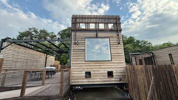 A wooden cabin with a deck, a small pond, and a fenced area.