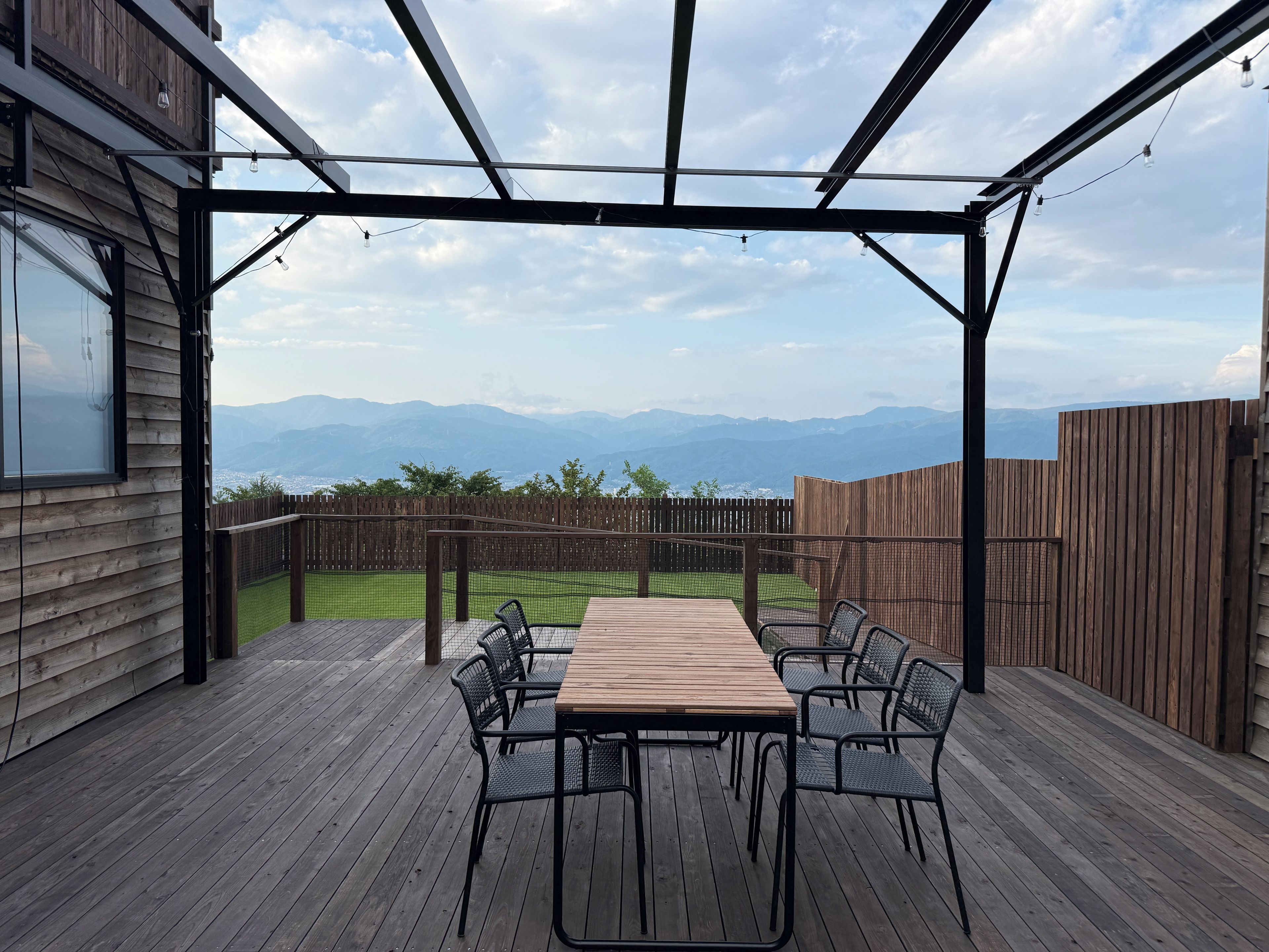 A covered outdoor patio with a wooden table and chairs, a view of mountains, and a wooden fence.