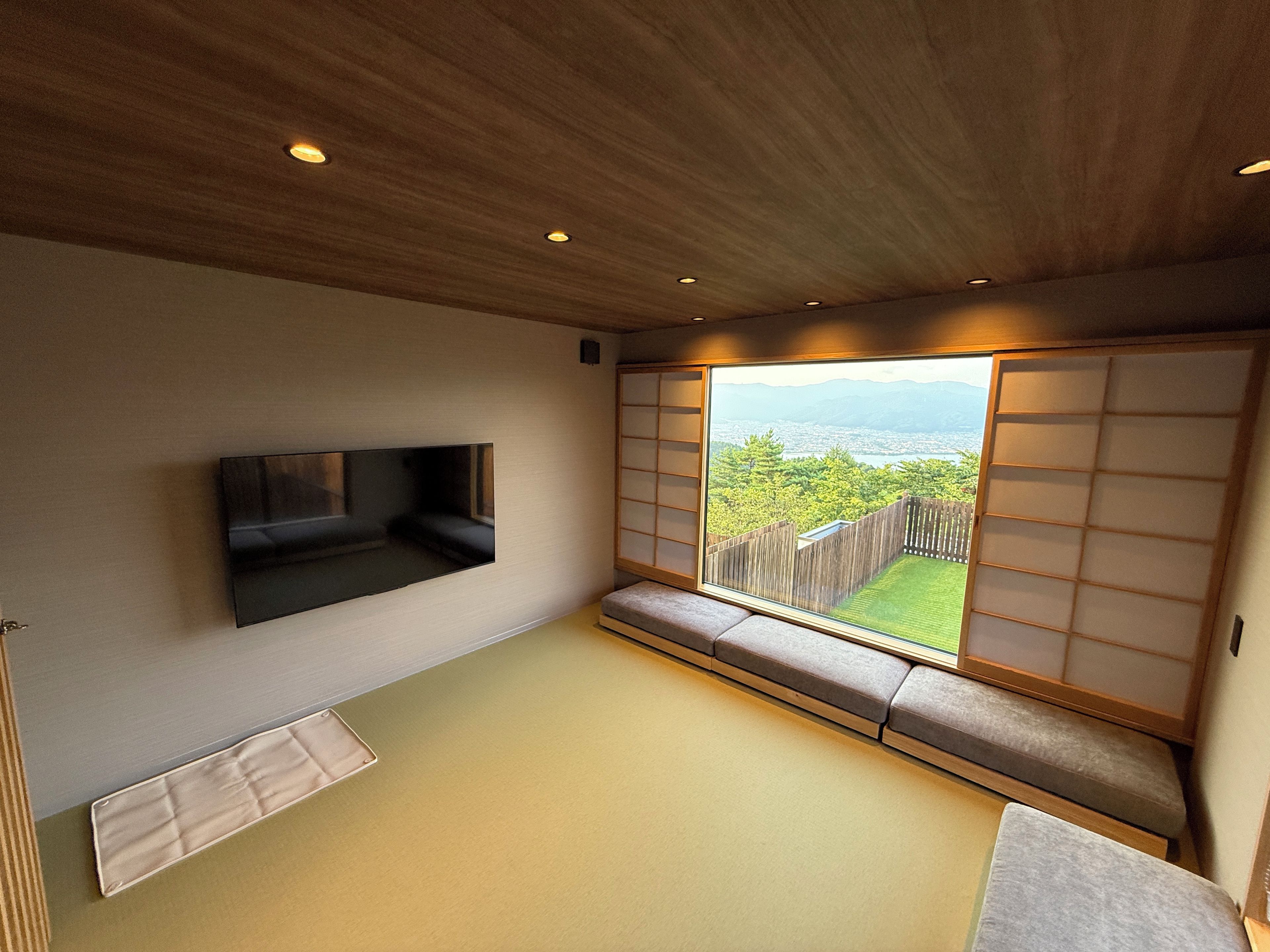 Una habitación con suelo de tatami, un televisor bajo montado en la pared, un panel corredero shoji y vistas a un paisaje verde.