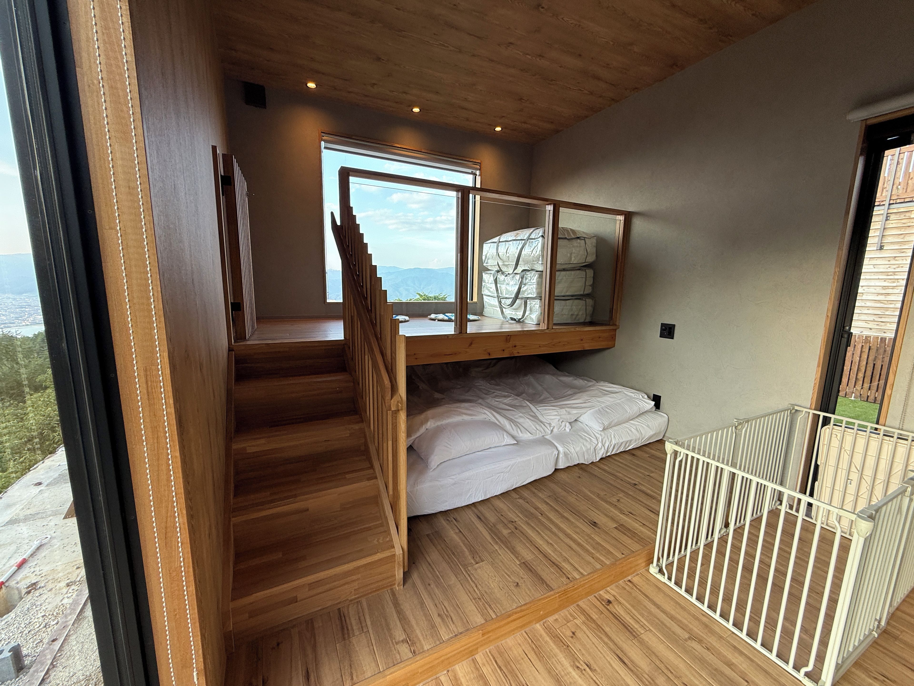 Une chambre avec un escalier en bois, un lit superposé, une fenêtre donnant sur un beau paysage et un balcon avec une balustrade blanche.
