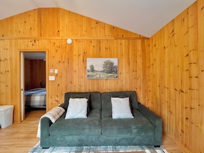 A room with wooden walls, a green sofa with two pillows, a framed landscape picture, and a door leading to another room. - Cozy Cottage #17 - King Bed -Pond View - Heated Pool (Brackley Beach)