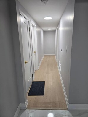 Um corredor estreito com piso de madeira, paredes brancas e um tapete preto na entrada.