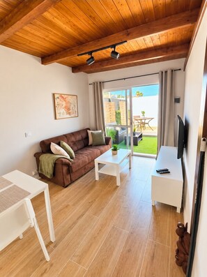 Ein möbliertes Wohnzimmer mit Holzboden, einer Couch, einem Couchtisch, einer TV-Bank und einer Glasschiebetür, die zu einem Außenbereich führt.