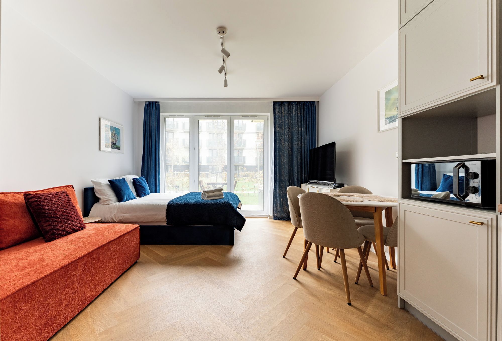 A modern hotel room with a bed, a sofa, a dining table, and a TV.