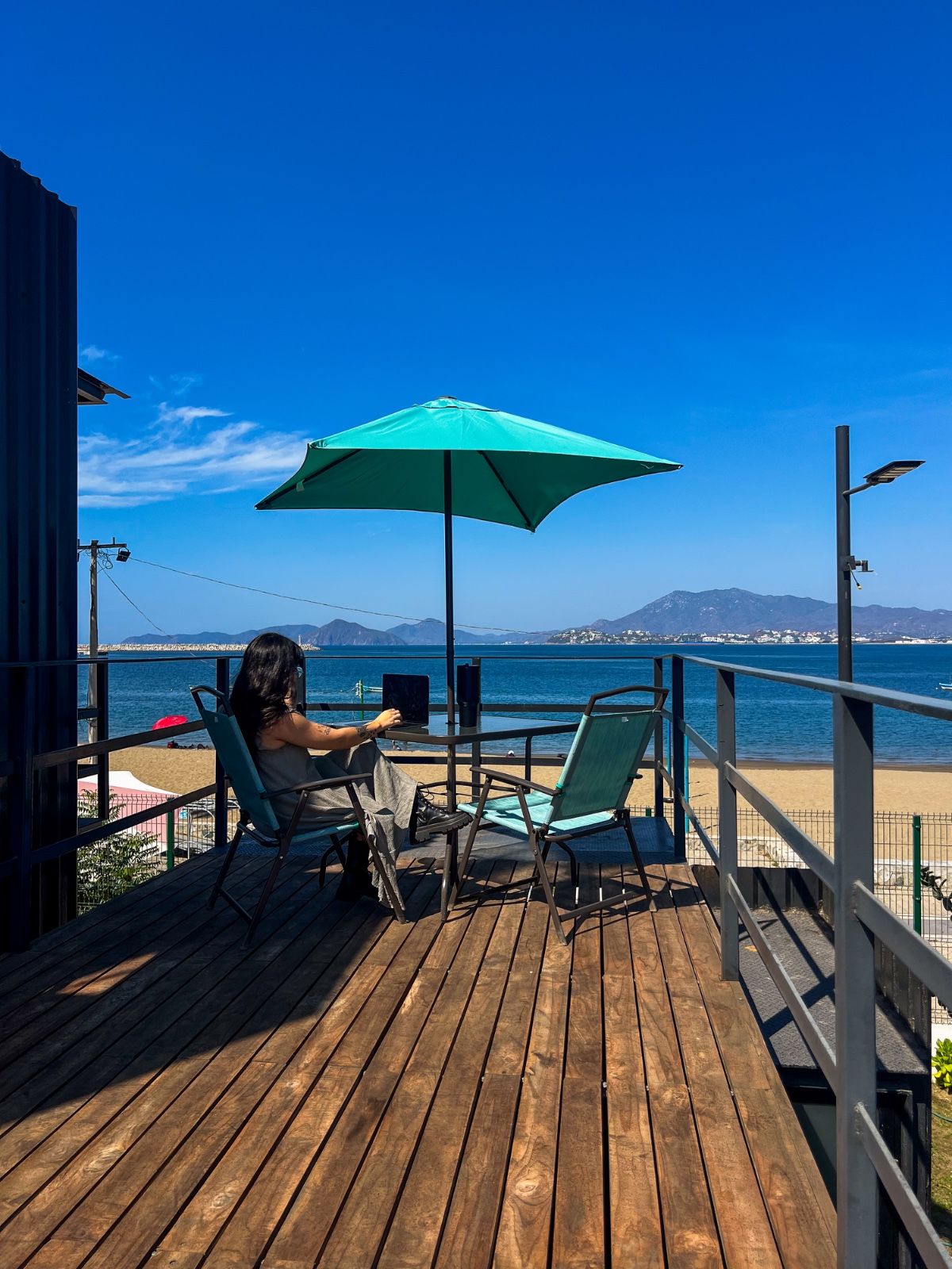 A wooden deck with two chairs and a table, overlooking a beach and the sea.