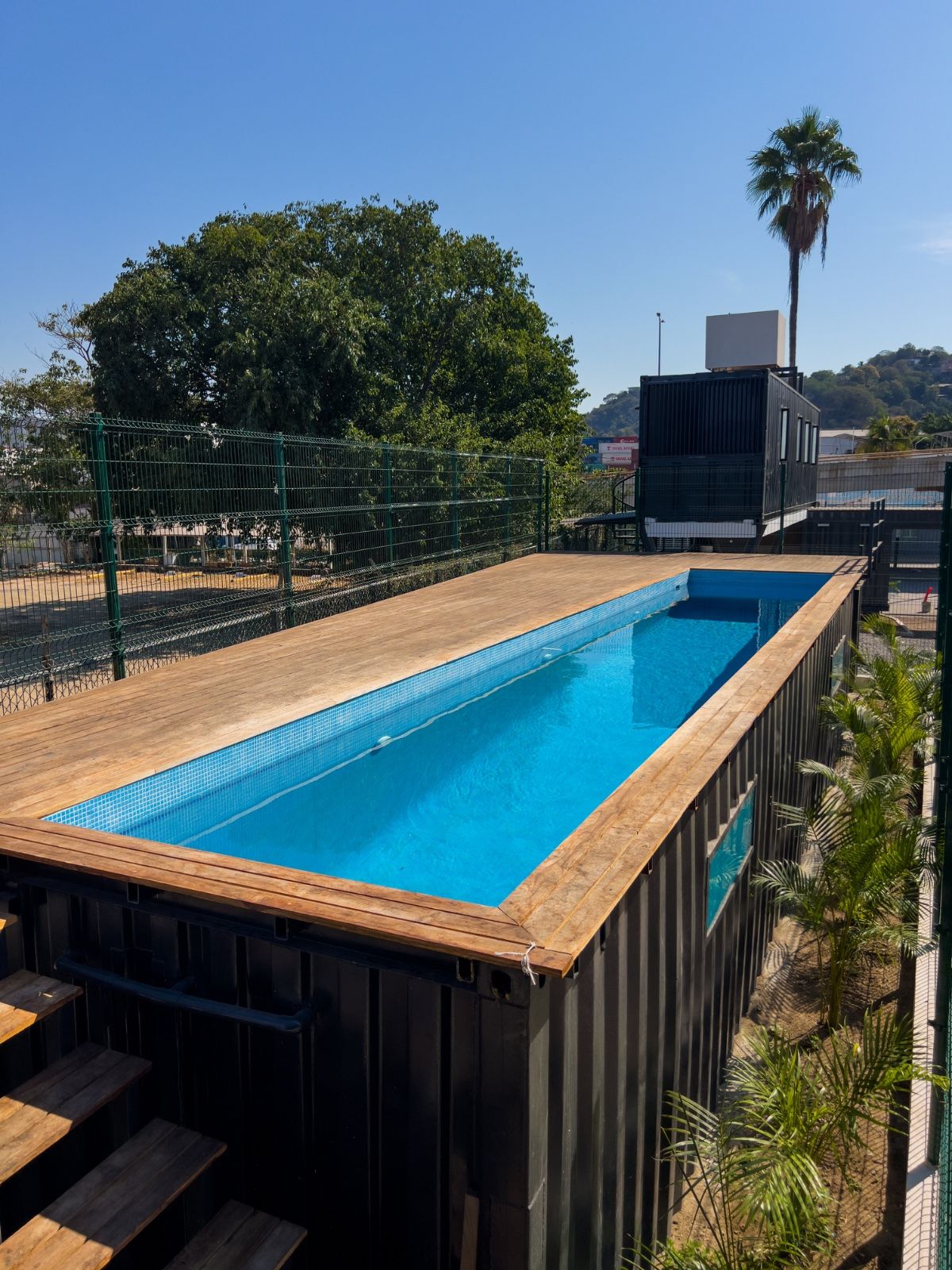 Piscina rectangular con borde de madera y rodeada por una cerca metálica, con árboles y un edificio al fondo.