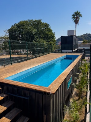 Une piscine rectangulaire bordée d’une terrasse en bois, entourée d’une clôture métallique, avec en arrière-plan des arbres et un bâtiment.
