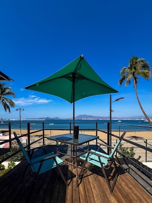 Une terrasse en bois avec une table et des chaises, un parasol vert et une vue sur la plage et l’océan.