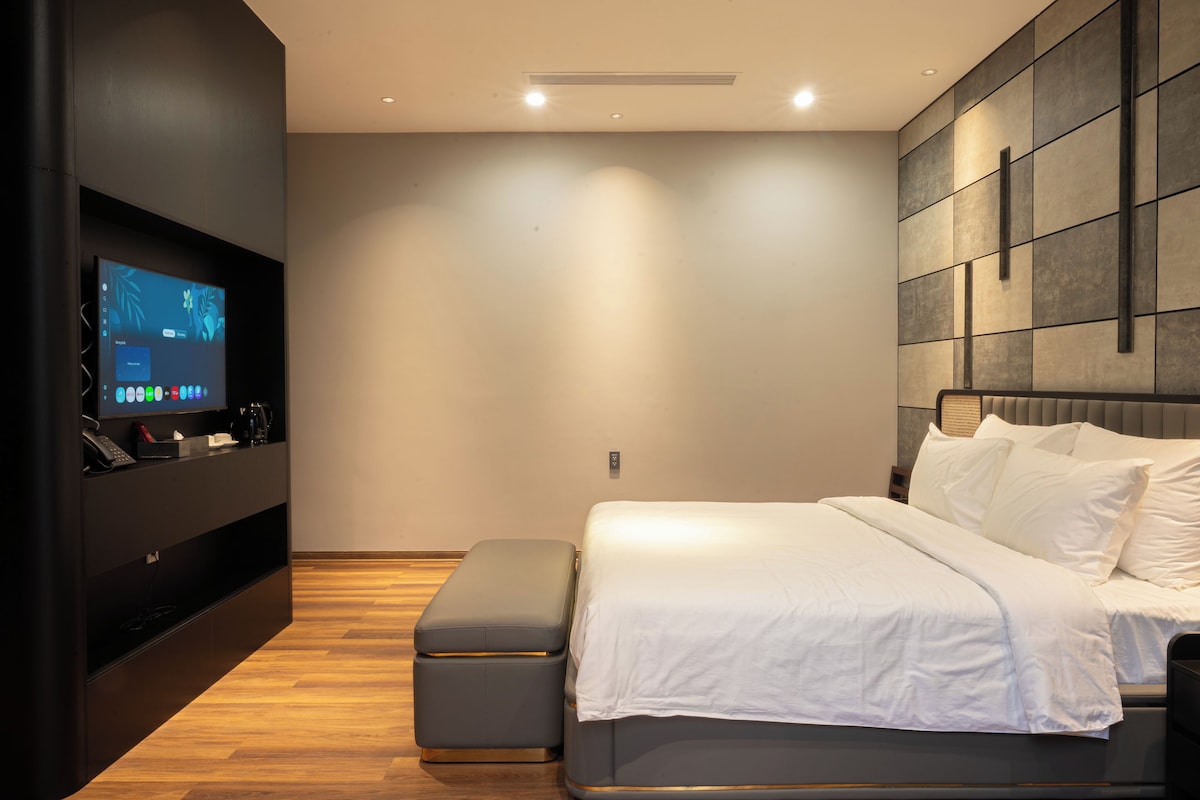 Ein modernes Schlafzimmer mit einem Bett, einem an einer dunklen Wand montierten Fernseher, einem Nachttisch mit Telefon und Lampe sowie einem Holzboden.