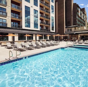 Ein Hotelpoolbereich mit Liegestühlen und Sonnenschirmen, umgeben von mehrstöckigen Gebäuden mit Balkonen.