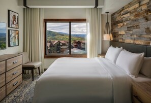 Ein Hotelzimmer mit einem großen Bett, einer hölzernen Kommode, einem Nachttisch, einer Lampe und Blick auf eine Stadt durch ein Fenster.