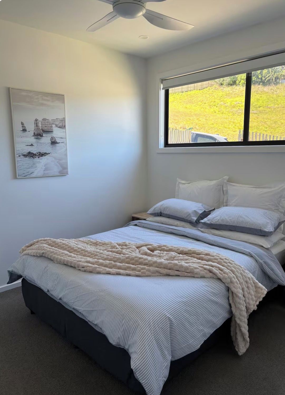 Une chambre à coucher avec un lit, un ventilateur de plafond, une fenêtre donnant sur un paysage verdoyant et un tableau accroché au mur.