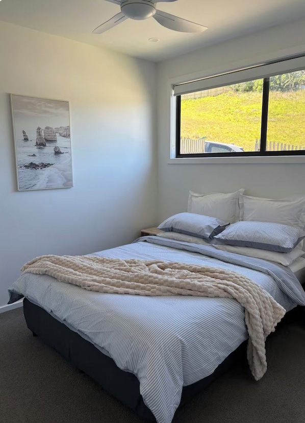 A bedroom with a bed, a ceiling fan, a window with a view of greenery, and a painting on the wall.