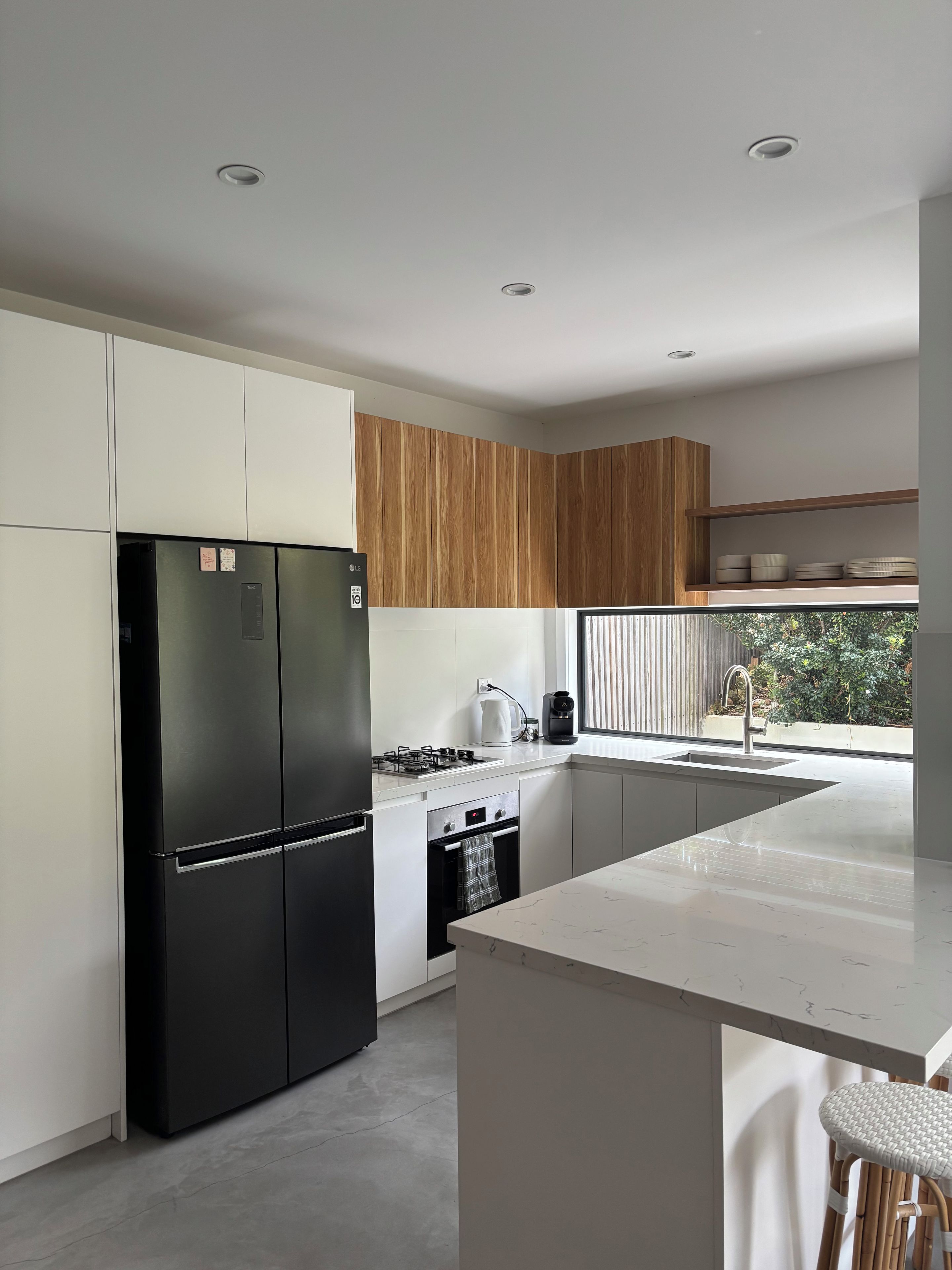 Une cuisine moderne avec un réfrigérateur noir, des armoires blanches, une armoire supérieure en bois, un four encastré et une fenêtre donnant sur un paysage verdoyant.