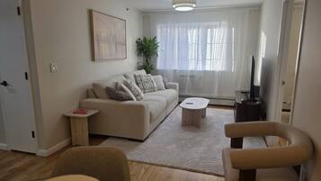 Ein Wohnzimmer mit einer Couch, einem Couchtisch und Sesseln. Ein Fernseher und eine Pflanze sind ebenfalls vorhanden.