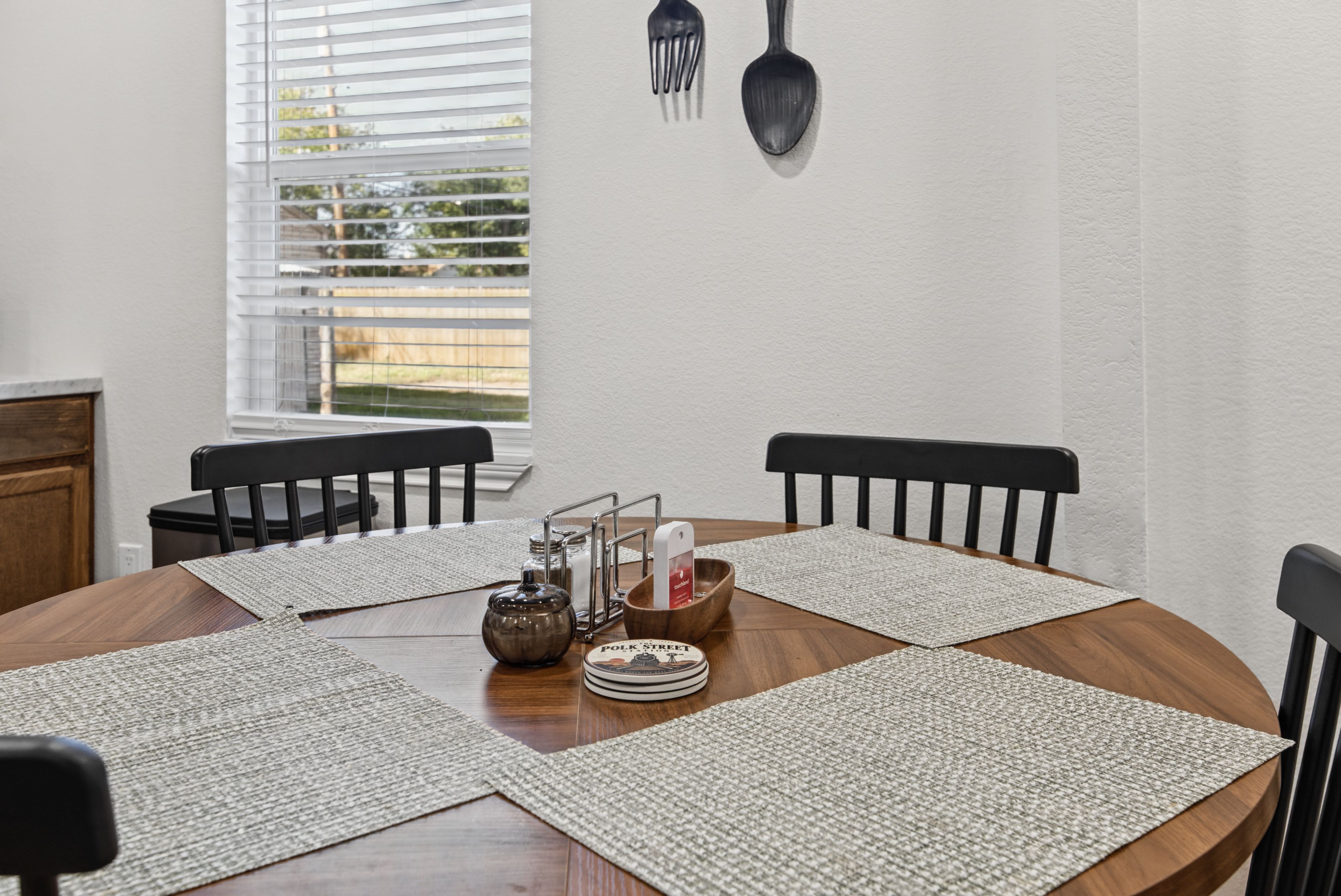Une table de salle à manger ronde, accompagnée de quatre chaises, une fenêtre avec des stores et un porte-ustensiles fixé au mur.