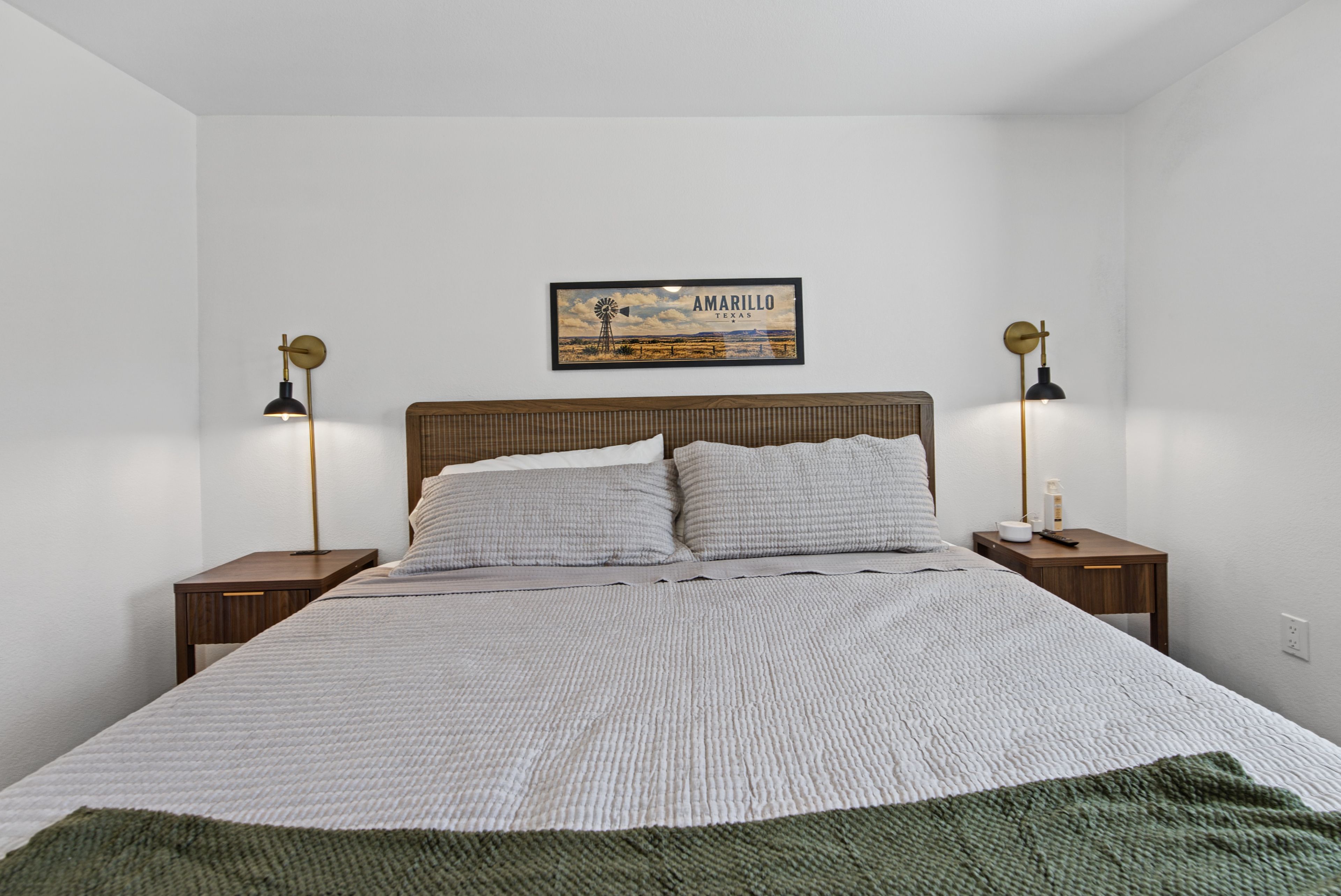 Une chambre à coucher avec un lit, deux tables de chevet, deux lampes, un tableau encadré au mur et une couverture verte sur le lit.