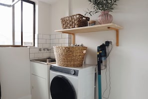 Un área de lavandería con lavadora, un cesto y un estante con una planta.