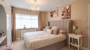 A bedroom with a large bed, a bedside table, a nightstand, a dresser, a window with curtains, and a chandelier. - 2-Bed House Sleeps 4 with Parking & Garden (Skegness)