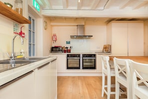 A modern kitchen with a stainless steel sink, double ovens, a microwave, and a wooden dining table with white chairs. - 11 Bedroom Cottage with Hot Tub and Parking (Derby)