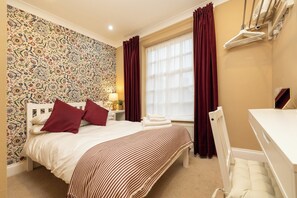 Ein Hotelzimmer mit Bett, Schreibtisch, Stuhl, einem fensterbepflanzten Fenster und einer Wand mit Blumenmuster.