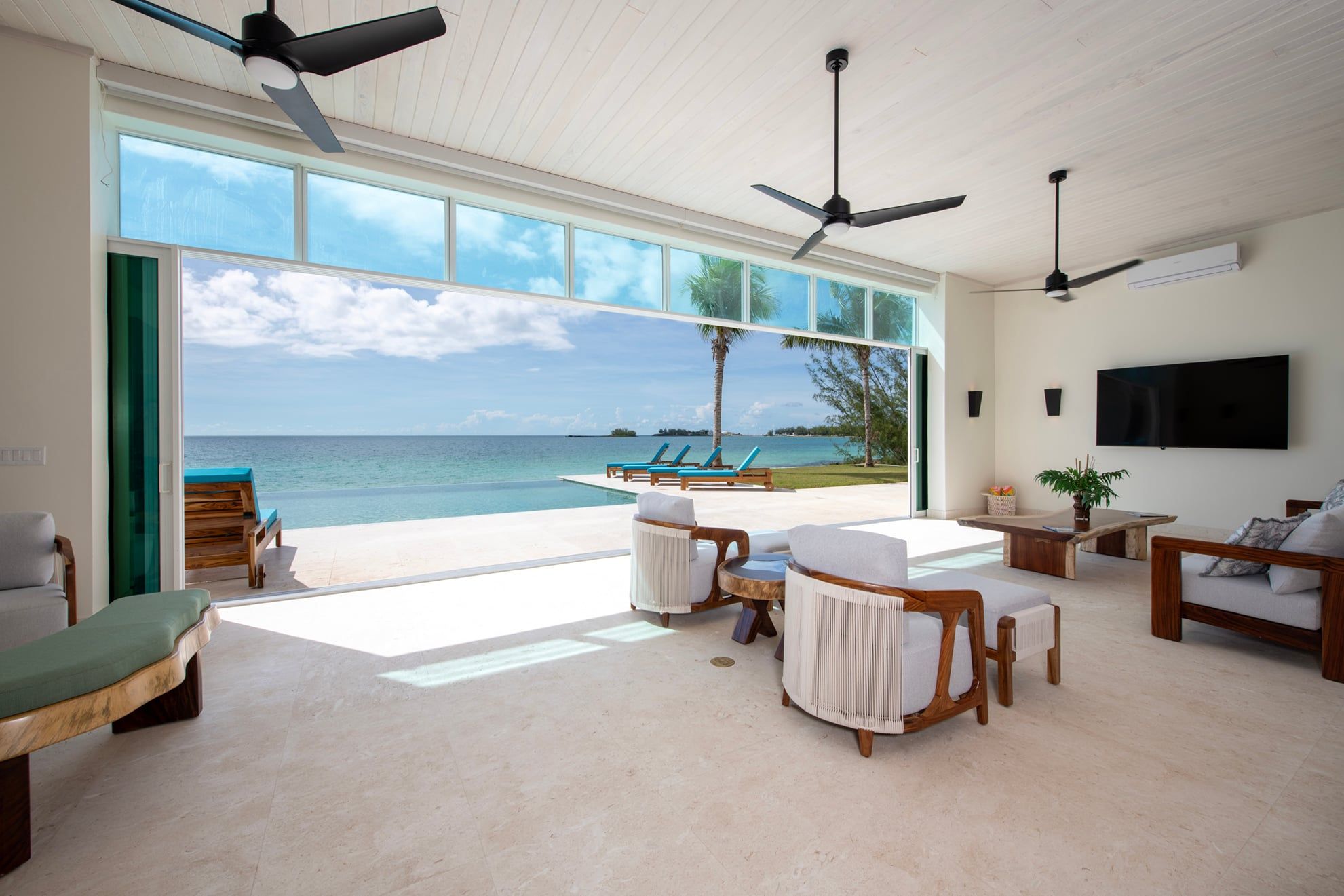Een ruime woonkamer met een groot raam met uitzicht op een strand, een plafondventilator en een flatscreen televisie aan de muur.