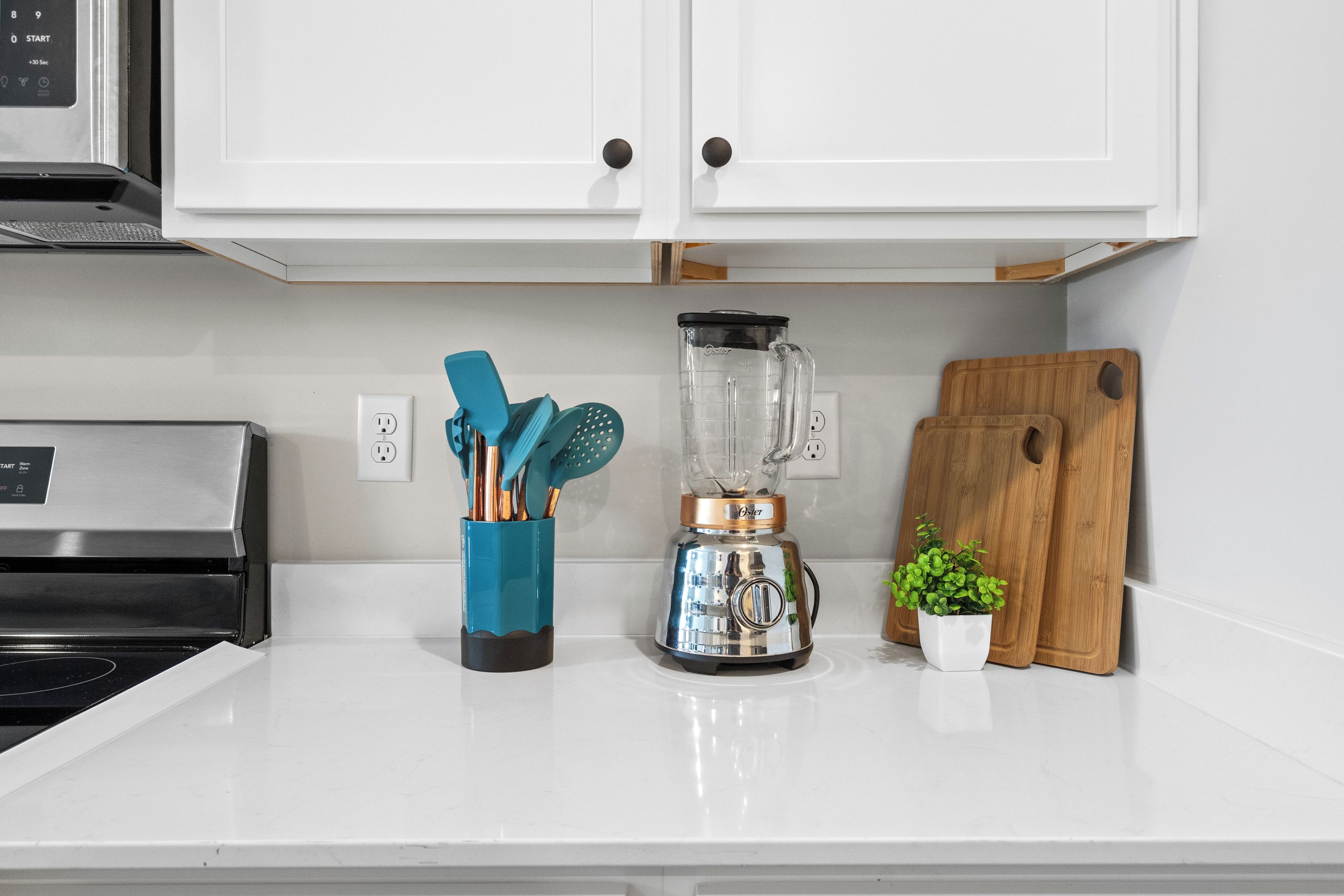 A kitchen with white cabinets, a stainless steel oven, a blender, a wooden cutting board, a plant, and a utensil holder.