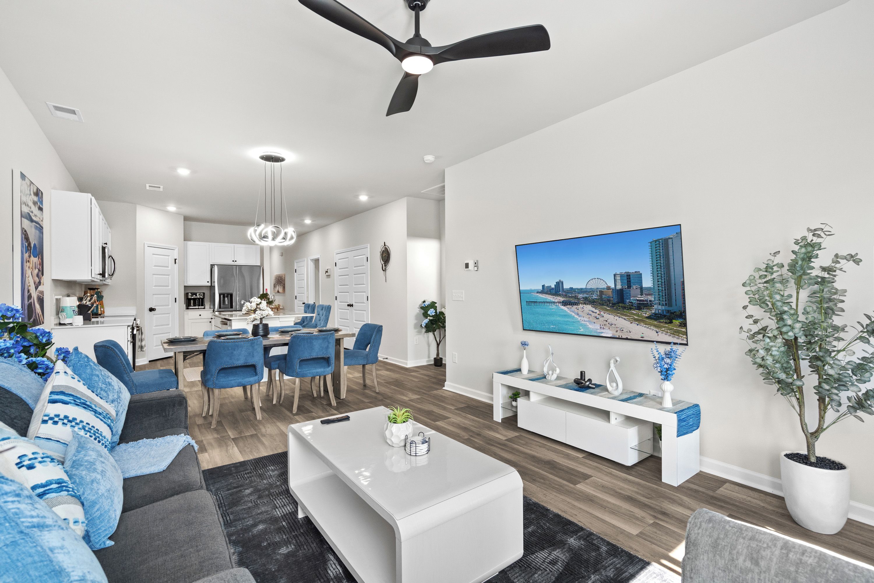A modern living room with a dining area, a ceiling fan, and a large flat-screen TV mounted on the wall.