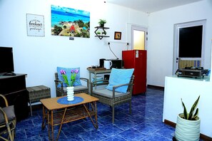 Un salon avec un téléviseur, une table à manger avec des chaises, un réfrigérateur rouge et une étagère murale ornée de plantes.