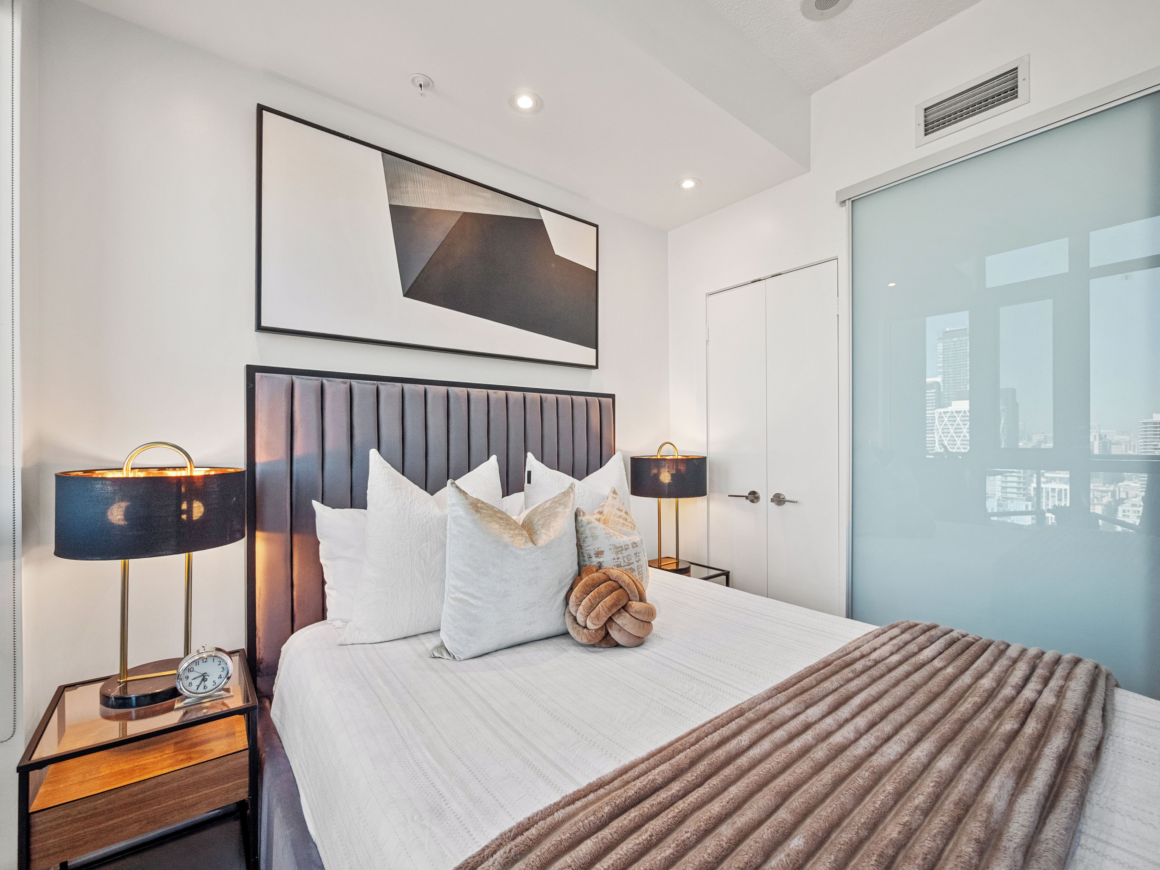 Een moderne hotelkamer met een groot bed, twee nachtkastjes, uitzicht over de stad en een ingelijst kunstwerk boven het bed.