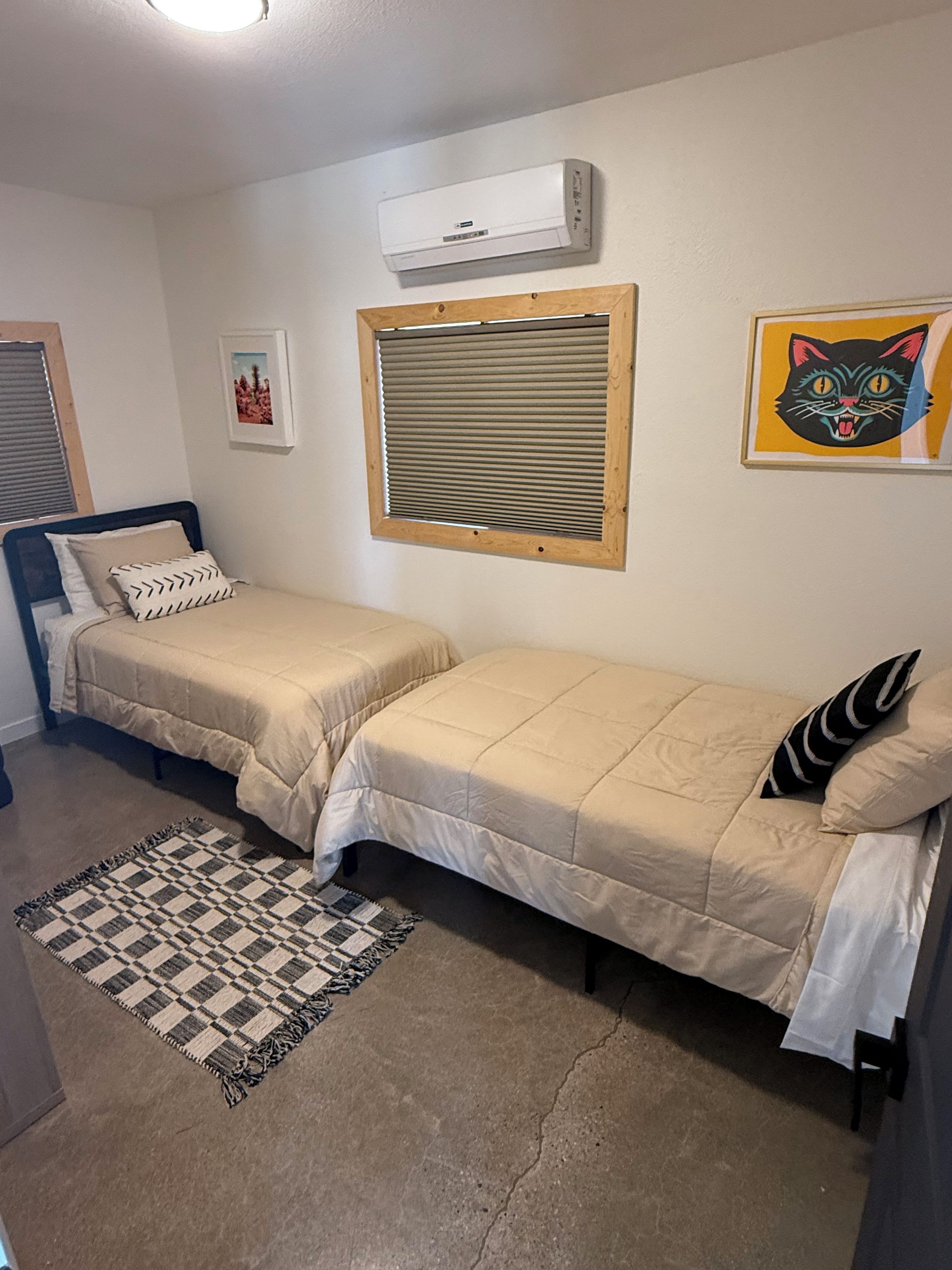 A bedroom with two beds, a window with blinds, a wall-mounted air conditioner, a framed picture, and a rug.