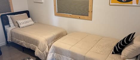 A bedroom with two beds, a window with blinds, a wall-mounted air conditioner, a framed picture, and a rug.
