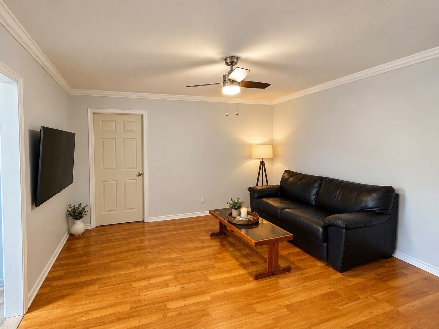 A living room with a black leather sofa, wooden coffee table, a ceiling fan, and a flat-screen TV mounted on the wall.