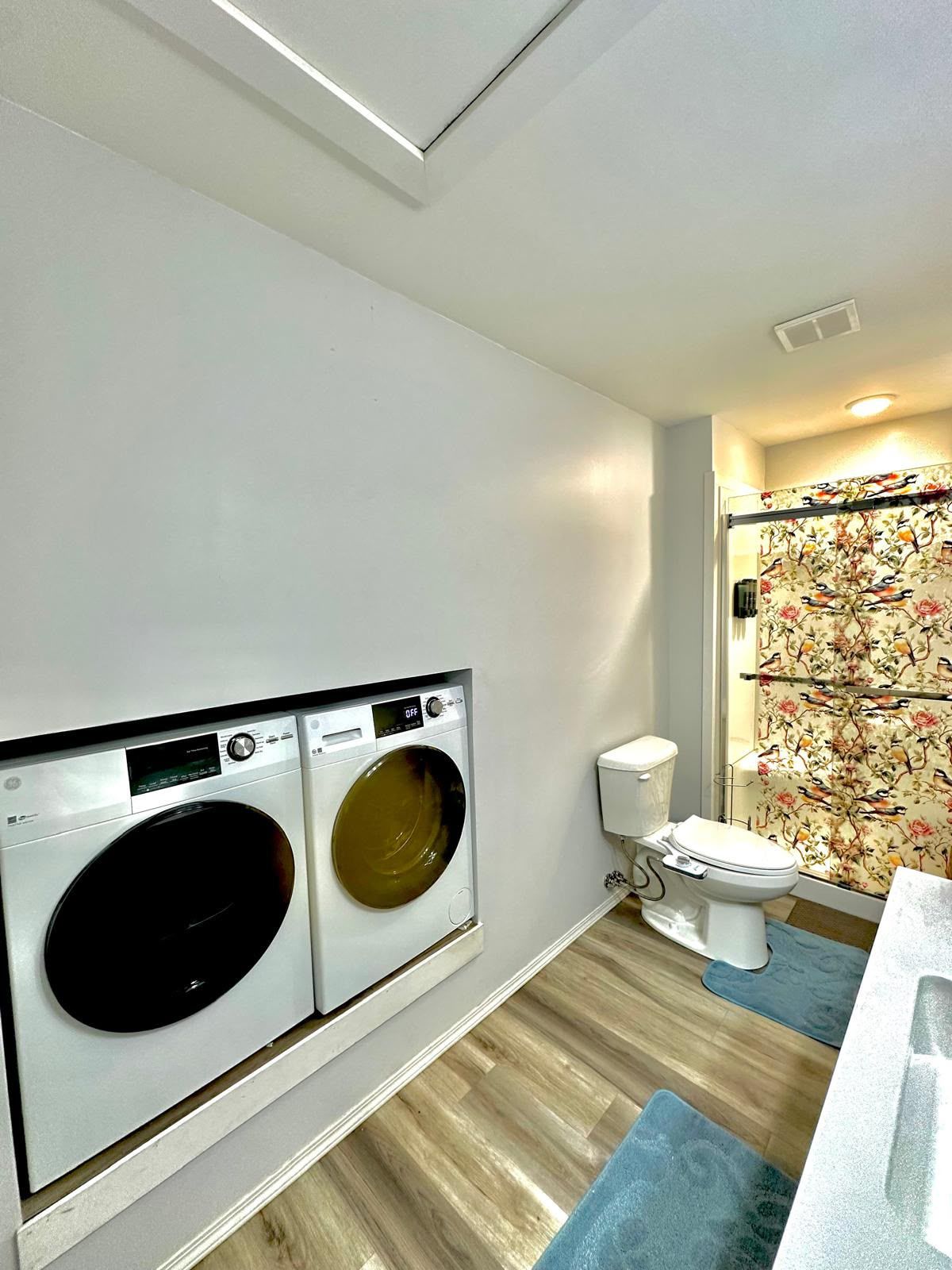 A laundry room with two front-loading washing machines, a toilet, and a shower with floral wallpaper.
