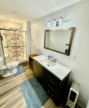 Une salle de bain moderne avec un lavabo blanc, des meubles sombres, un grand miroir et une douche agrémentée d’un rideau de douche à motifs floraux.