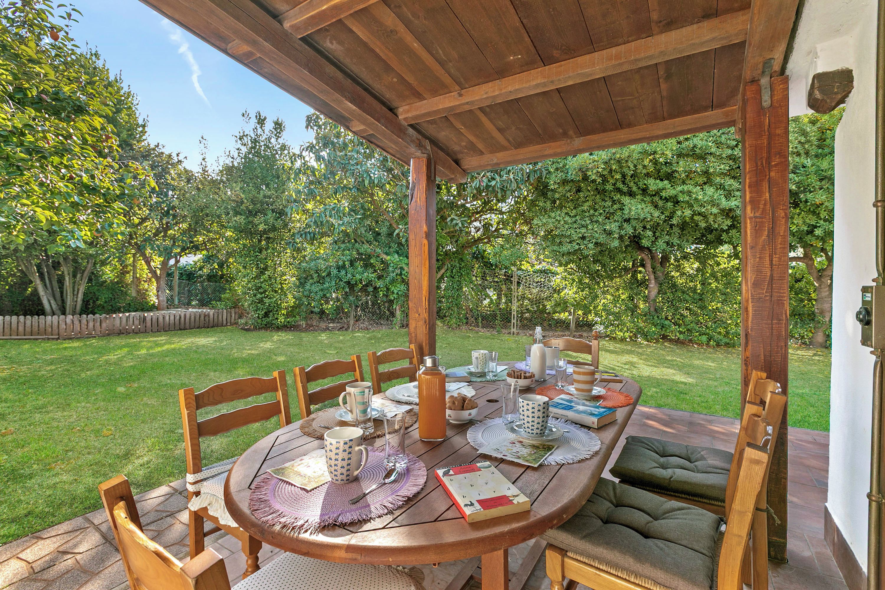 An outdoor wooden pergola with a round table set for dining, surrounded by chairs, and a garden view.