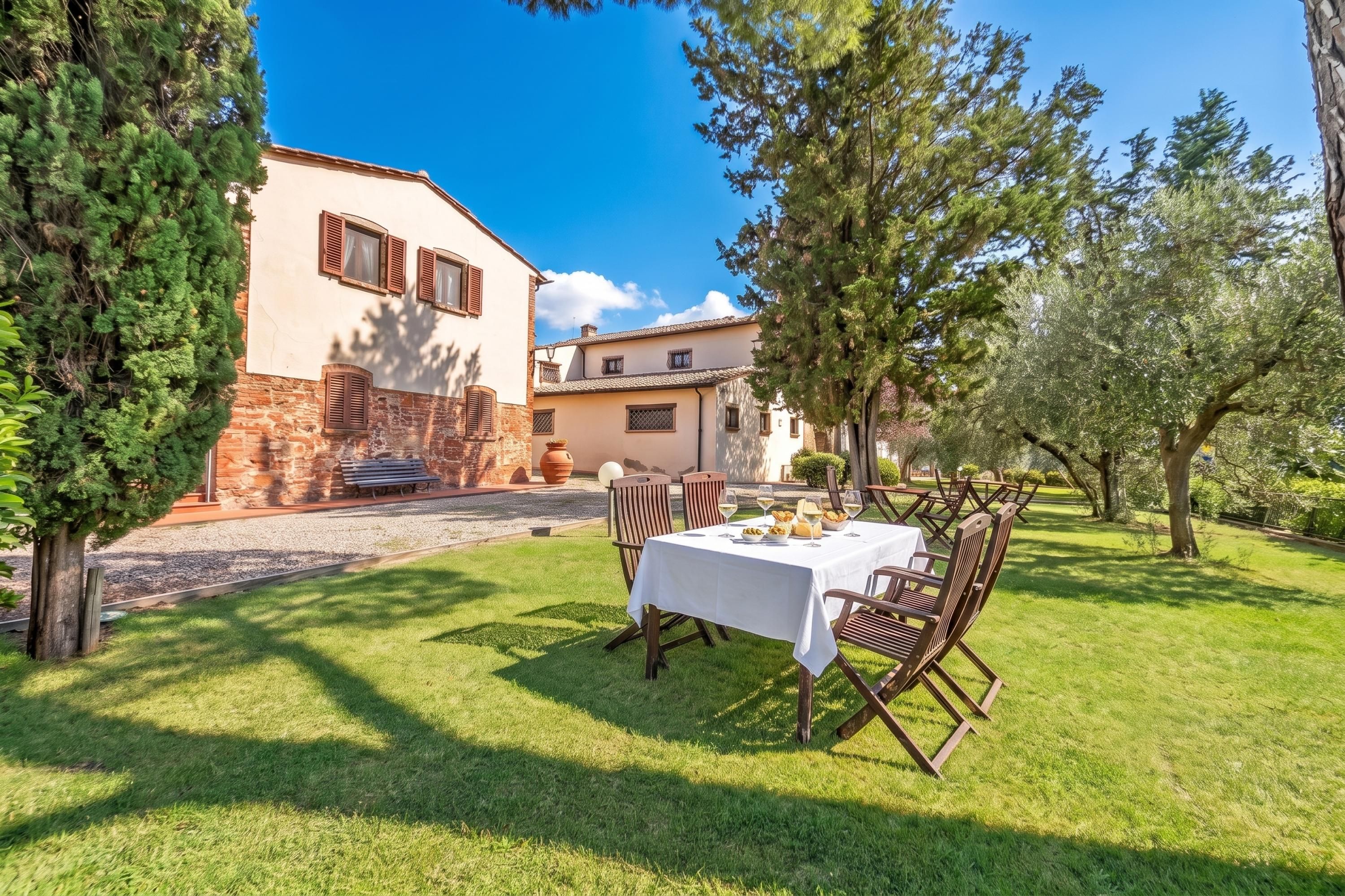 A dining area with a table set for two, surrounded by chairs, in a garden with a building in the background.