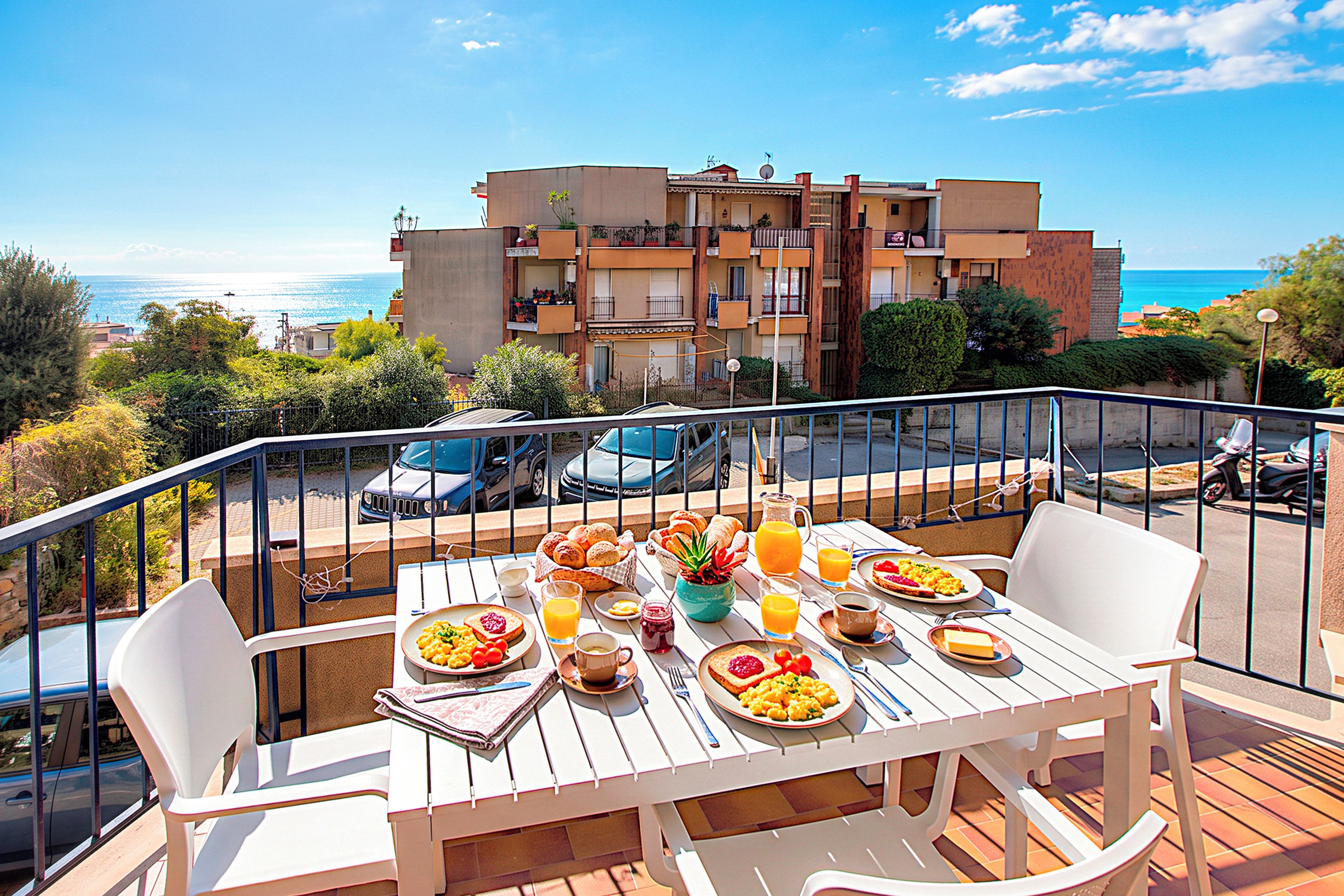 A balcony with a table set for breakfast, overlooking a coastal area with buildings and parked cars.