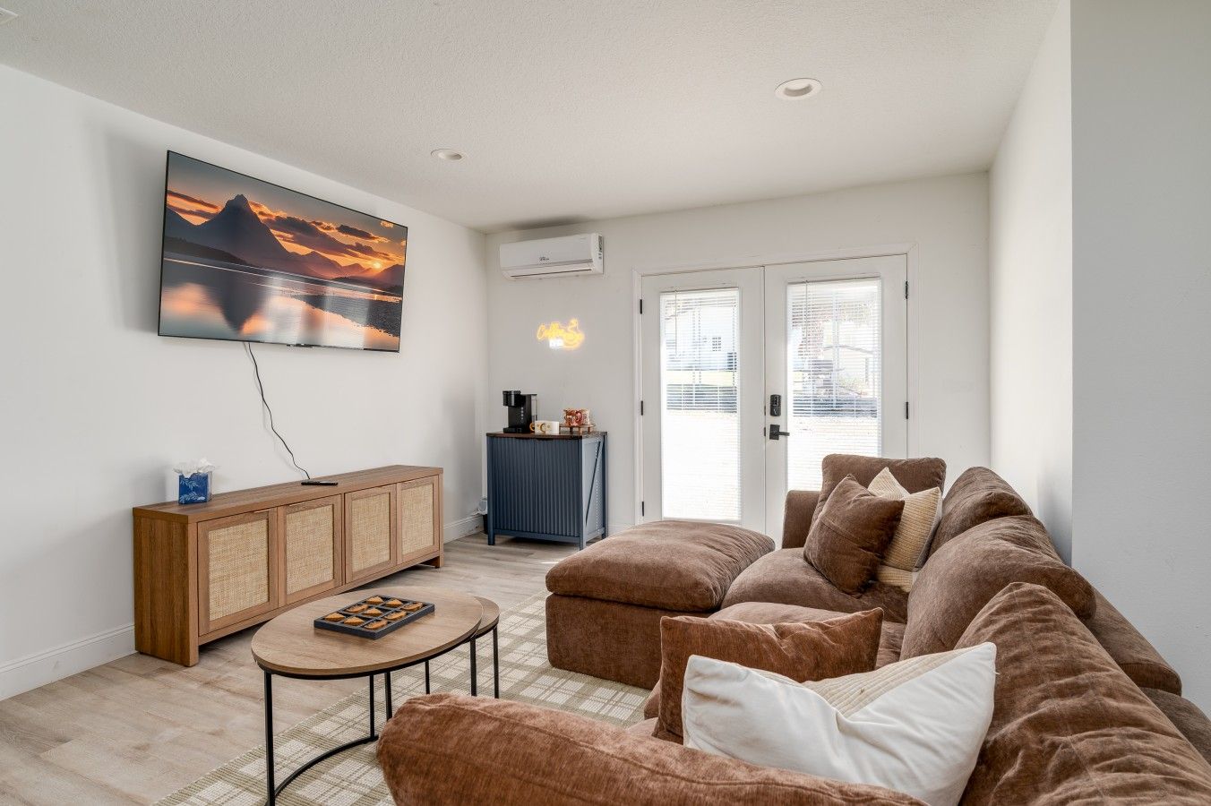 A living room with a brown sofa, a coffee table, a TV mounted on the wall, a cabinet, and a door leading outside.