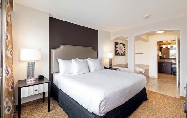 Habitación de hotel con una cama grande, mesitas de noche, un escritorio y vistas al baño y la cocina.
