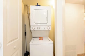 Un lavadero pequeño con lavadora y secadora apiladas, un trapeador y una puerta que da a otra habitación.