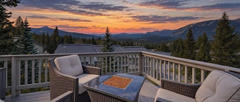 A deck with wicker furniture and a fire pit, overlooking a mountainous landscape.