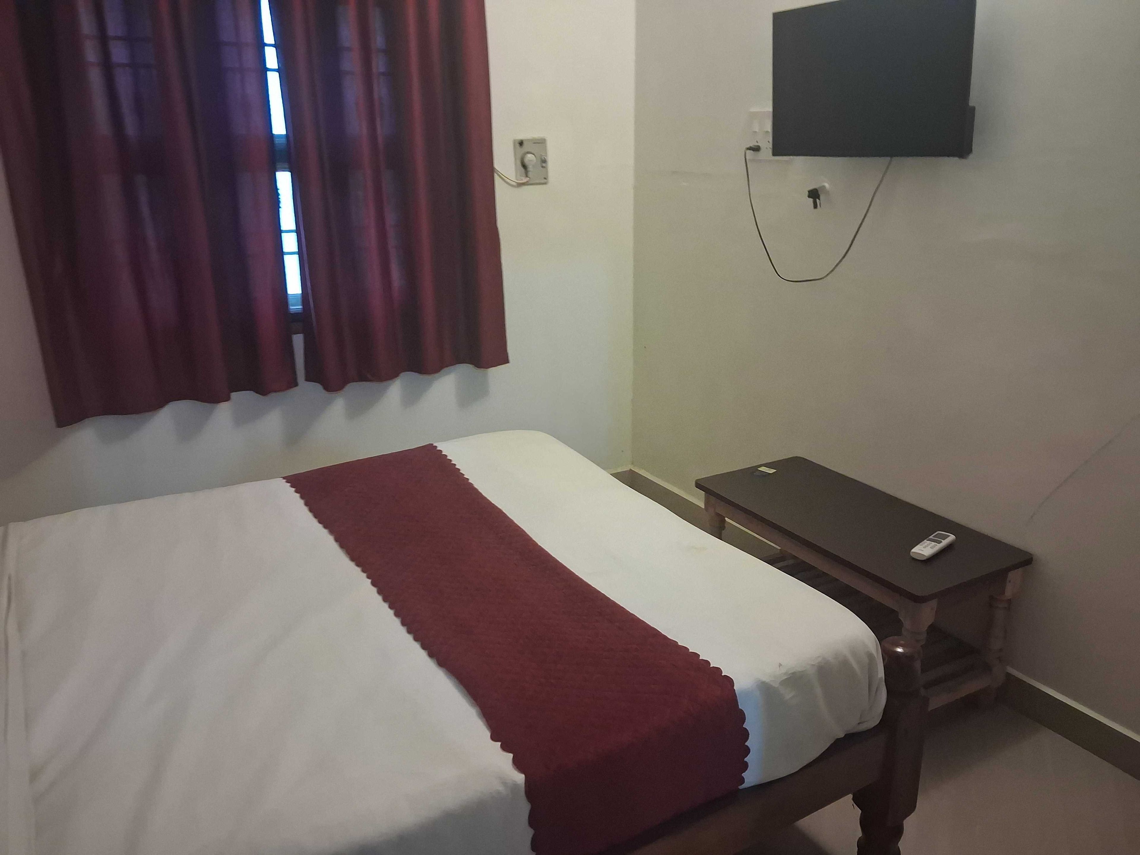 Una cama individual con una colcha blanca y roja, una mesita de noche pequeña, un televisor montado en la pared y una ventana con cortinas.