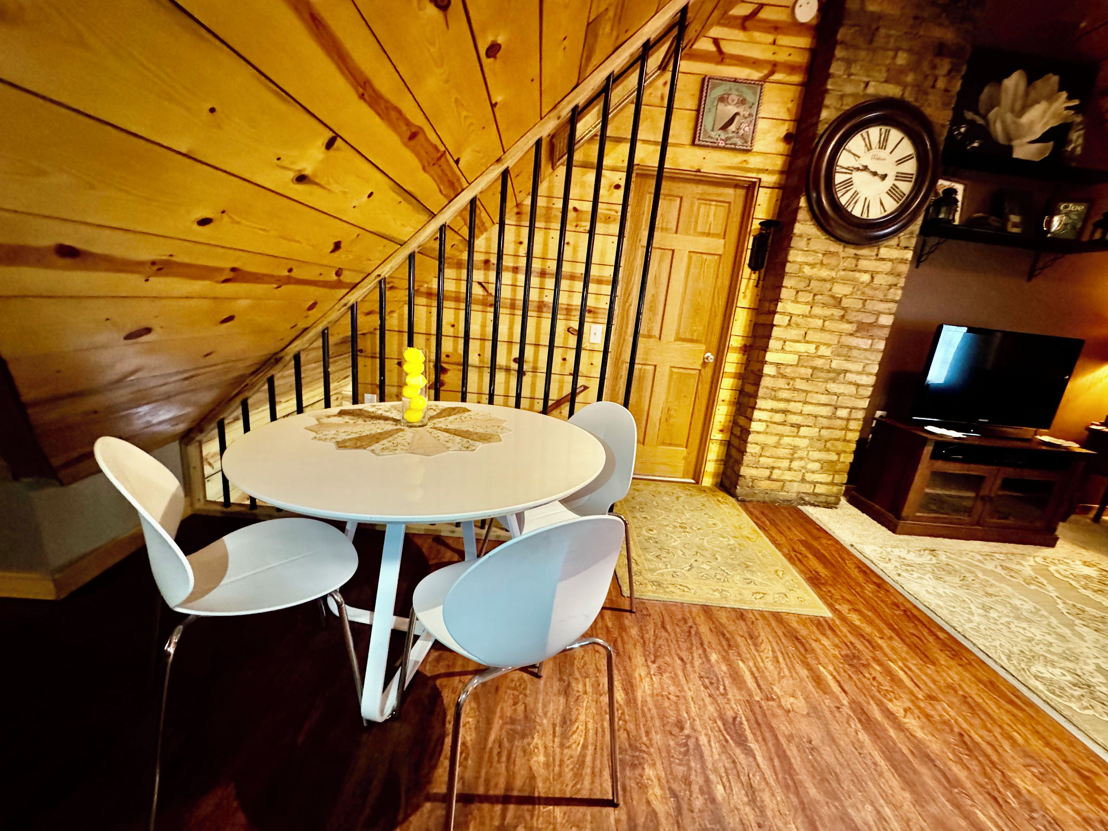 A dining area with a round table and chairs, a wooden staircase, a brick wall, and a television on a wooden stand.
