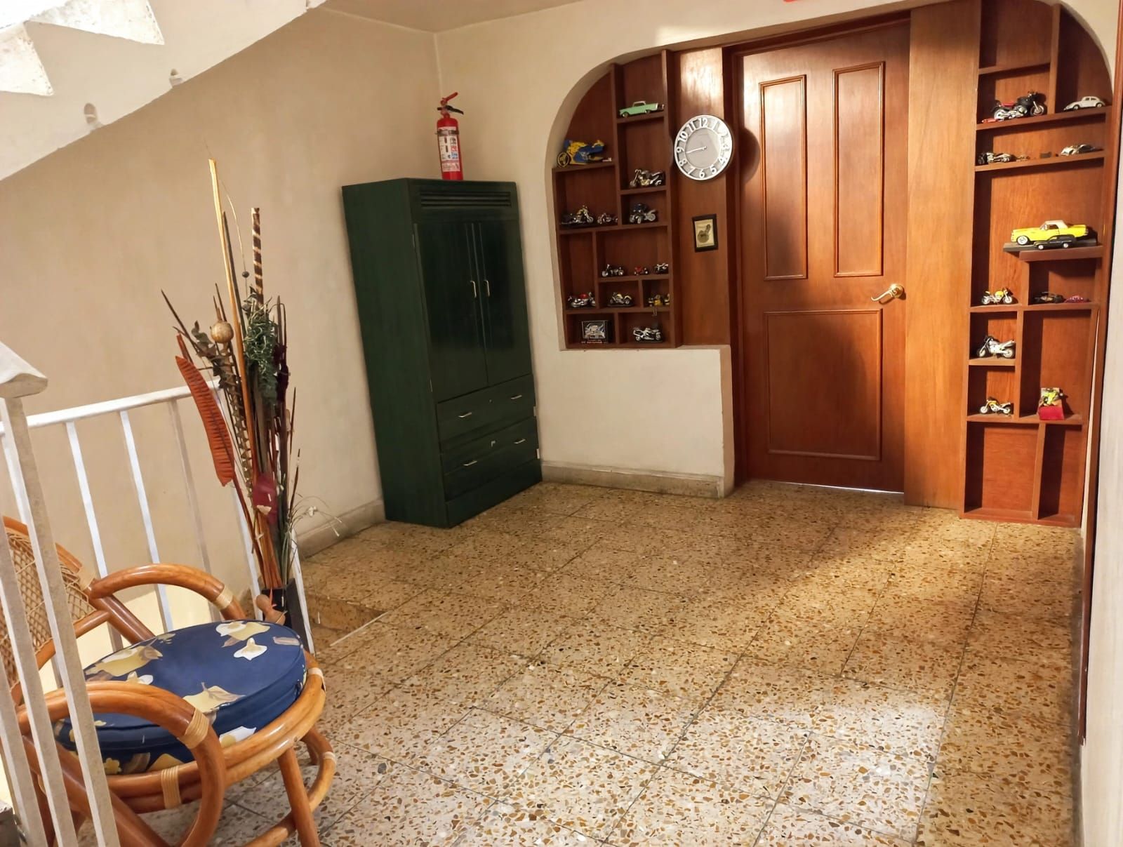 Una habitación con puerta de madera, un mueble verde, una silla con cojín azul, y una pared con un reloj y estantes llenos de juguetes.