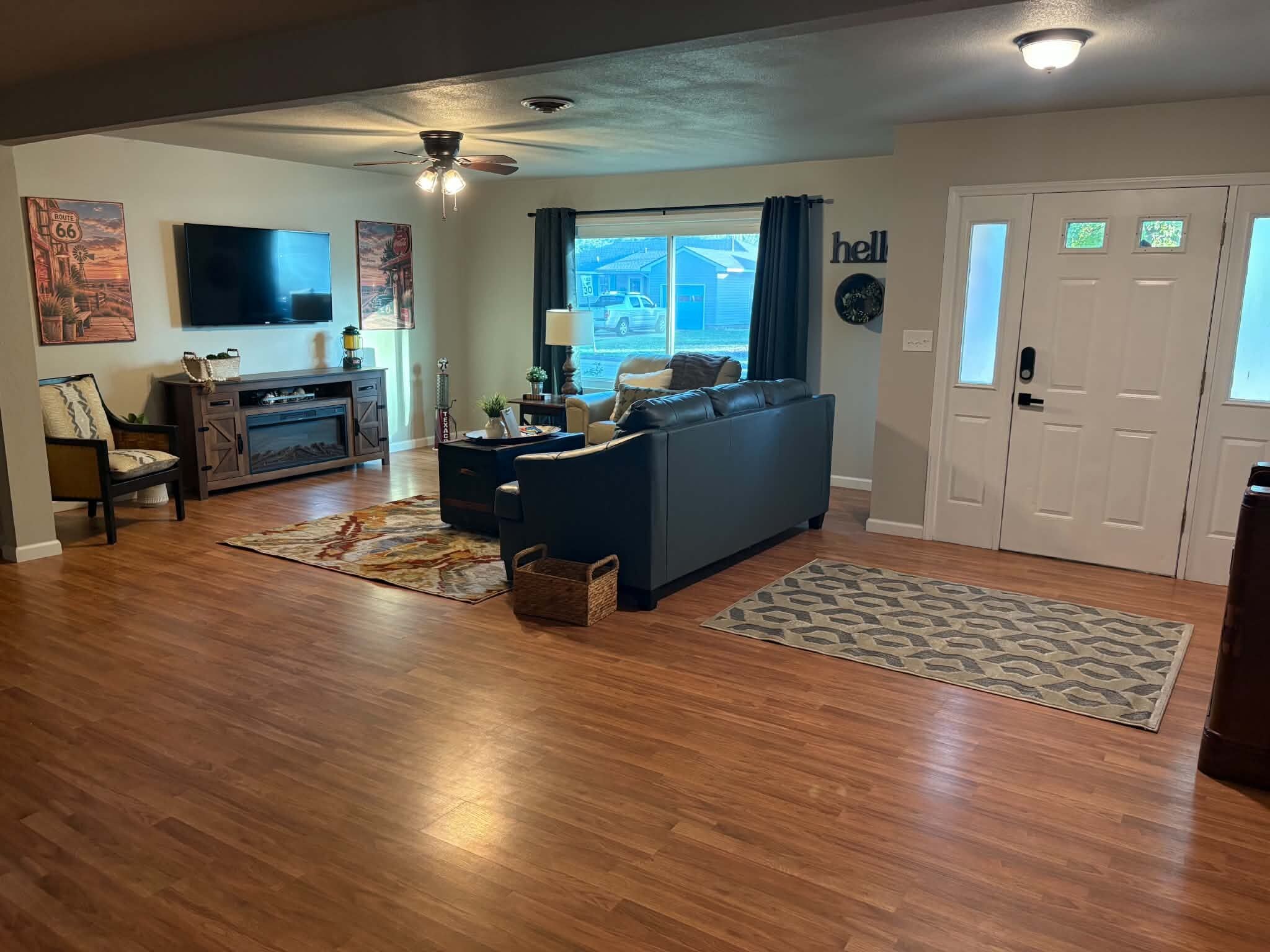 A spacious living room with hardwood flooring, a dark blue sofa, a wooden entertainment unit, and a large window with curtains.