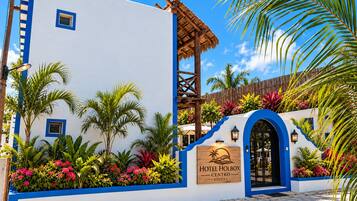 A hotel entrance with a blue and white facade, a wooden signboard, and tropical plants.