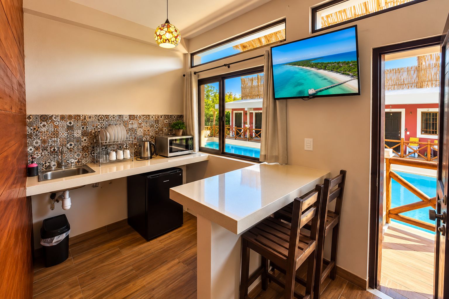 A modern kitchen with a large island, a dining area with wooden chairs, a television mounted on the wall, and a view of a pool through the windows.