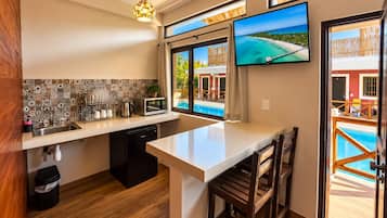 A modern kitchen with a large island, a dining area with wooden chairs, a television mounted on the wall, and a view of a pool through the windows.